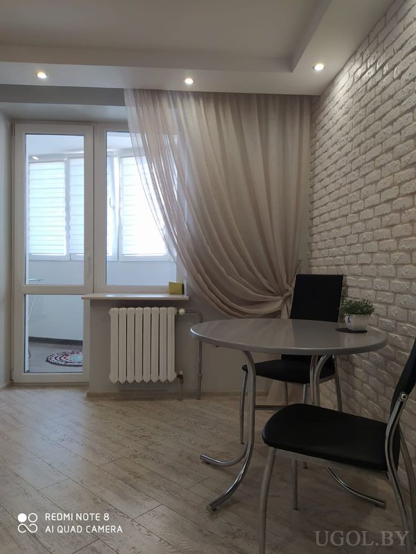 Небольшая столовая с круглым столом на металлической ножке, двумя чёрными стульями, белой кирпичной стеной и выходом на балкон с жалюзи.