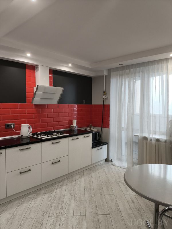 Современная кухня с белыми шкафами, чёрной столешницей, красным фартуком из плитки и встроенной техникой.