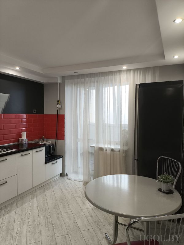 Минималистичная кухня с белыми шкафами, красным фартуком, круглым столом с металлическими стульями и высоким чёрным холодильником.