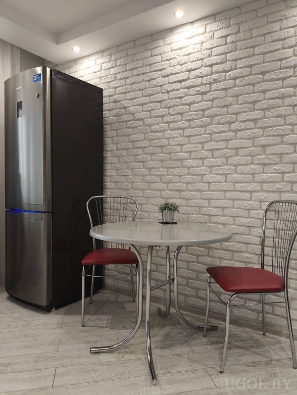 Компактная кухня с круглым столом, металлическими стульями с красными сиденьями и высоким холодильником у белой кирпичной стены.