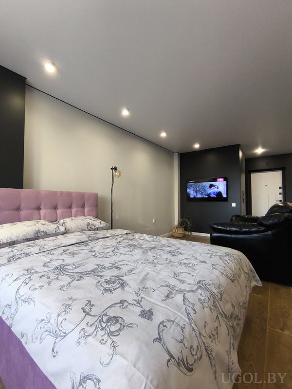 Современная спальня с кроватью в белом покрывале с серым узором, мягким сиреневым изголовьем, чёрным диваном и телевизором на стене.