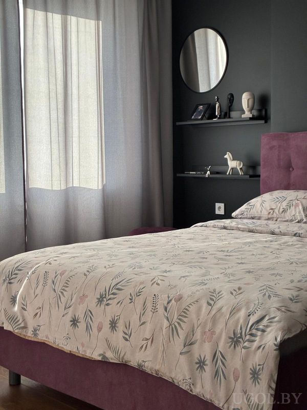 Уютная спальня с кроватью в светлом покрывале с узором, сиреневым изголовьем, тёмной стеной с полками и декоративными предметами.