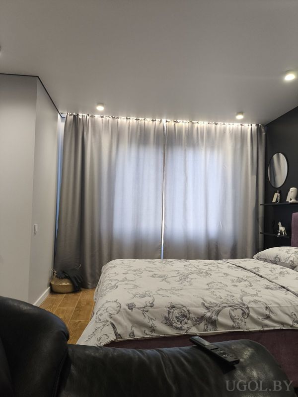 Современная спальня с кроватью в сером покрывале с узором, тёмной акцентной стеной с зеркалом и креслом с пультом на переднем плане.