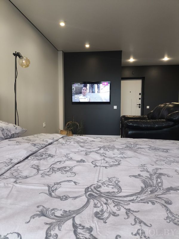 Минималистичная спальня с кроватью в сером покрывале, телевизором на тёмной стене и чёрным диваном сбоку.