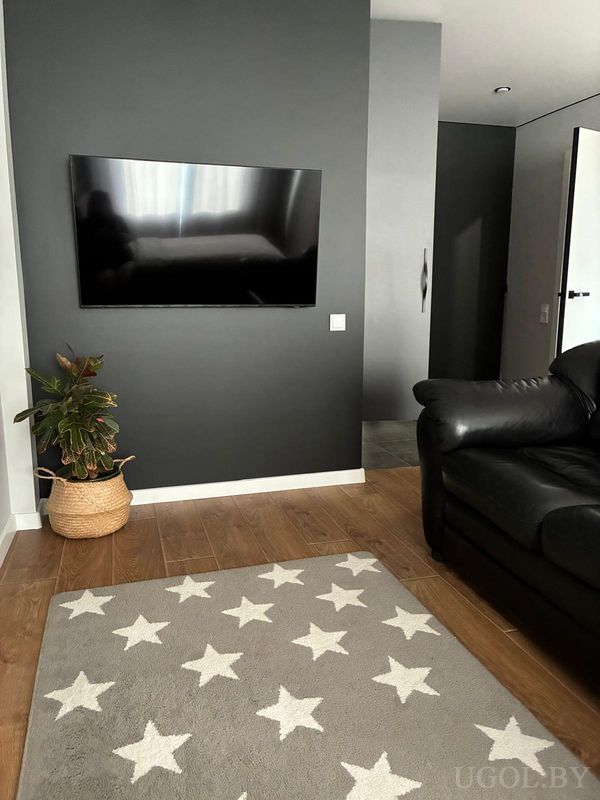 Современная гостиная с телевизором на тёмной стене, ковром со звёздами, чёрным диваном и деревянным полом.