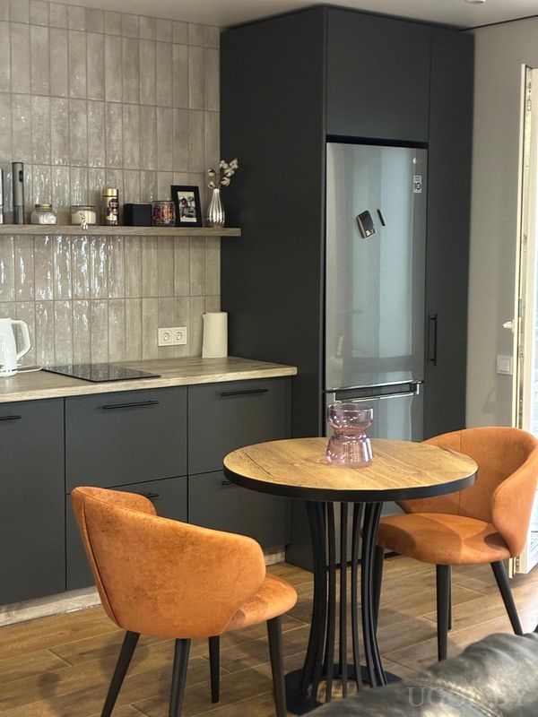 Современная кухня с серыми шкафами, плитой, чайником, деревянной столешницей и круглым столом с оранжевыми креслами.