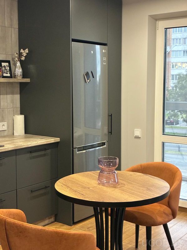 Кухня с серыми шкафами, встроенным холодильником, круглым столом с оранжевыми креслами и видом на жилой квартал из окна.
