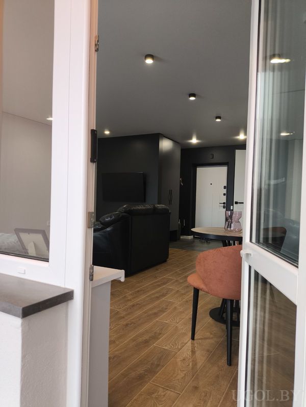 Современный интерьер с персиковым креслом, чёрным диваном, телевизором на тёмной стене и деревянным полом.