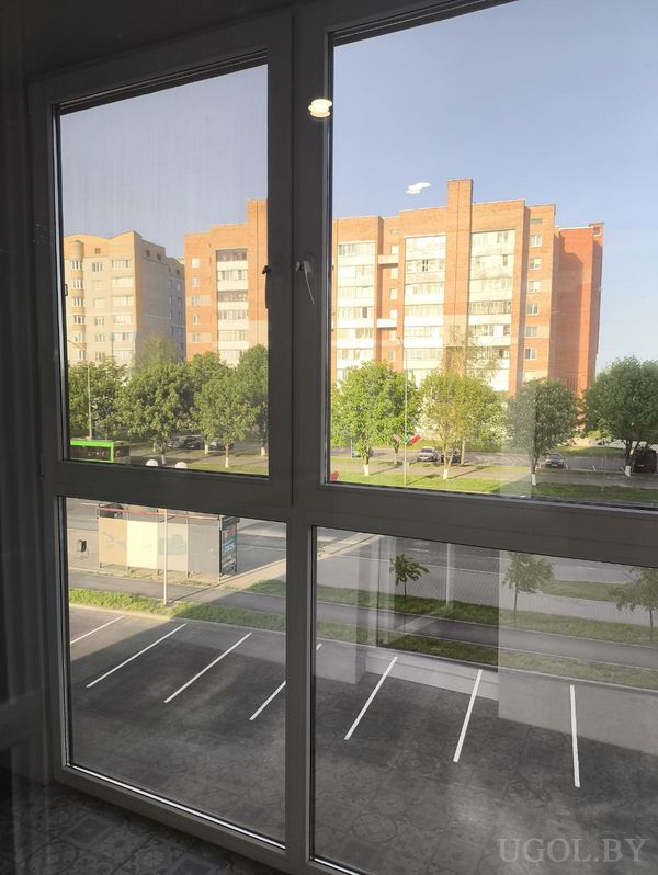 Городской вид из окна на улицу с припаркованными машинами, зелёным автобусом и жилыми домами в солнечную погоду.