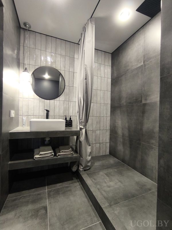 Минималистичная ванная с белым умывальником, круглым зеркалом, серой плиткой и подвесным светильником.