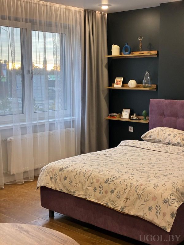 Современная спальня с кроватью в цветочном покрывале, мягким фиолетовым изголовьем и тёмной стеной с тремя деревянными полками и декором.