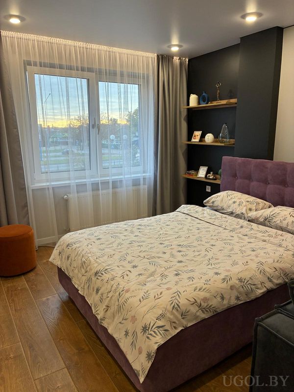 Уютная спальня с кроватью в светлом покрывале с растительным узором, фиолетовым изголовьем, тёмной стеной с полками и оранжевым пуфом у окна.