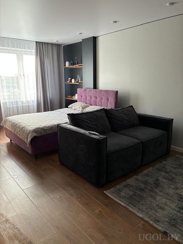 Современная спальня с кроватью в светлом покрывале, фиолетовым изголовьем, серыми шторами и тёмным диваном на ковре.