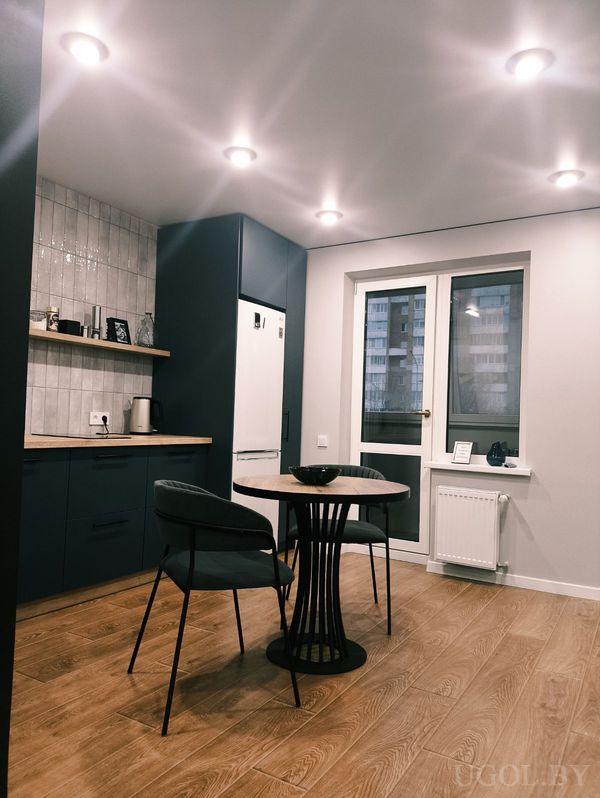 Компактная кухня с тёмно‑синими шкафами, плиткой на фартуке, деревянной полкой и круглым столом с двумя тёмными креслами.