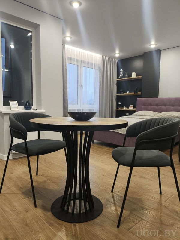 Интерьер студии с круглым столом и двумя креслами, фиолетовой кроватью у окна и чёрным стеллажом с декором.
