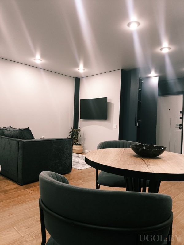 Современная гостиная с круглым столом и четырьмя креслами, тёмным диваном и телевизором на стене.