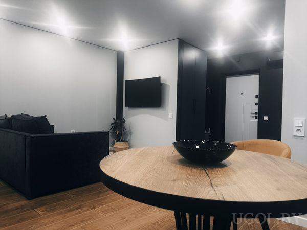 Минималистичная гостиная с круглым столом, тёмным диваном, телевизором на стене и серыми стенами.