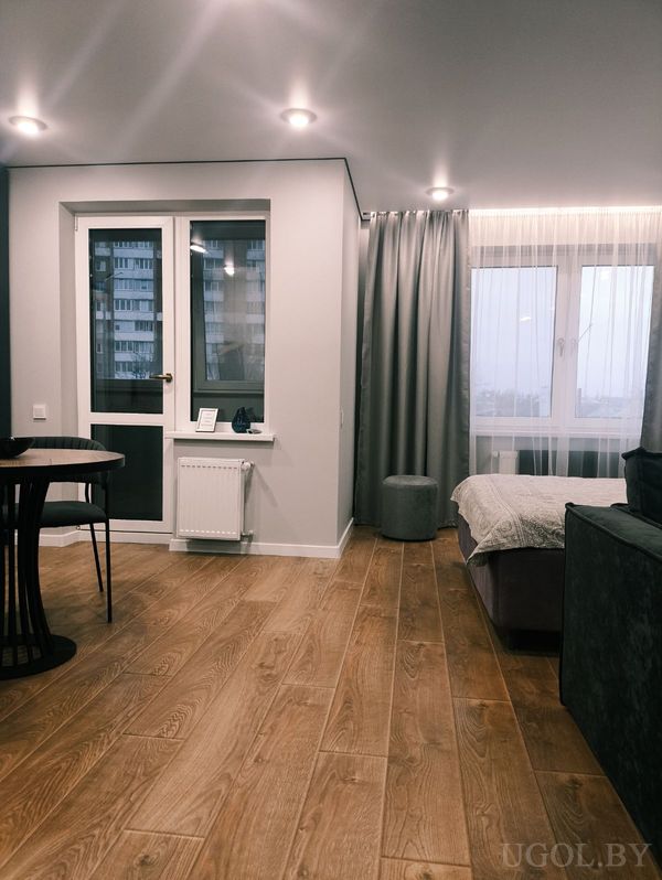 Уютная студия с кроватью в светлом покрывале, круглым столом с двумя креслами и серыми шторами у окна.