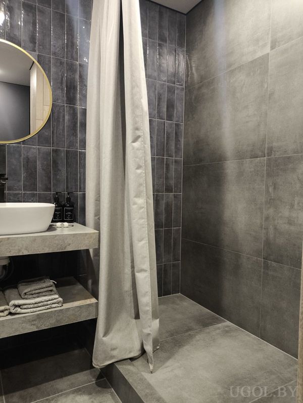 Минималистичная ванная с белым умывальником, круглым зеркалом в золотой оправе и открытой полкой с полотенцами.