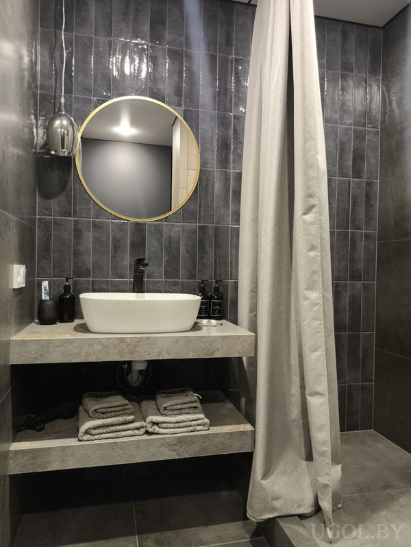 Современная ванная с тёмной плиткой, белым умывальником, круглым зеркалом в золотой оправе и открытой полкой с полотенцами.