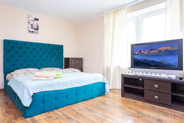 Светлая спальня с кроватью в бирюзовом изголовье, прикроватными тумбами и телевизором на тумбе.