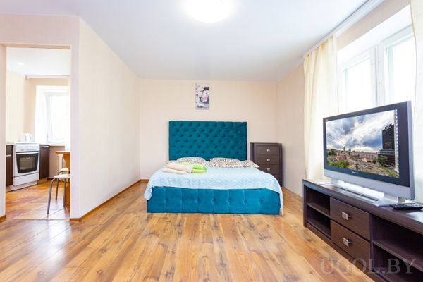 Современная студия с кроватью в бирюзовом изголовье, телевизором, кухней и обеденным столом.