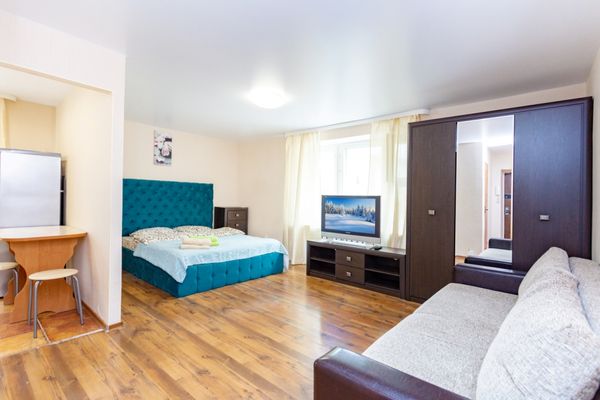 Студия с кроватью, диваном, телевизором, шкафом с зеркалами и обеденным столом у холодильника.