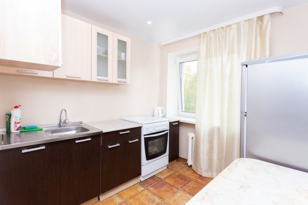 Небольшая кухня с плитой, мойкой, белыми и коричневыми шкафами и окном с бежевыми шторами.