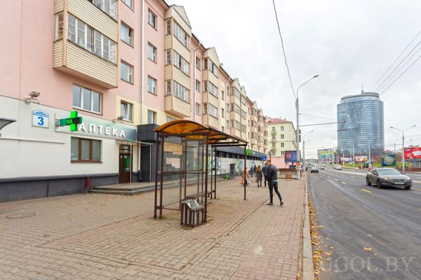 Городская улица с автобусной остановкой, аптекой на первом этаже дома и высоким стеклянным зданием на фоне.