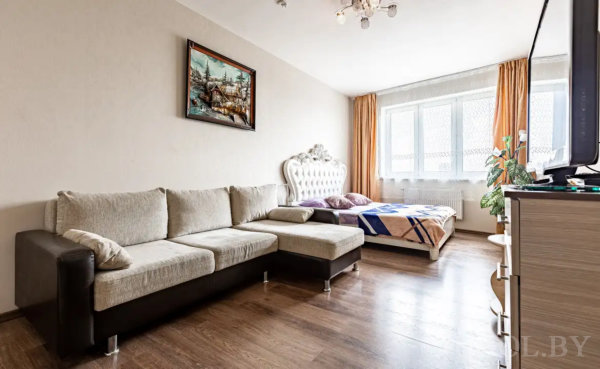 Жилая зона с диваном, кроватью с фигурным изголовьем, телевизором, оранжевыми шторами и растением у окна.