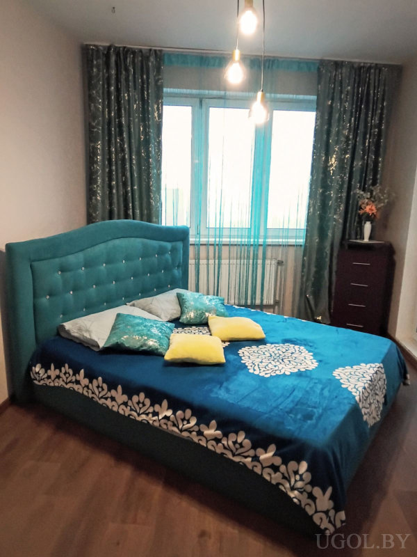 Интерьер с кроватью, бирюзовыми шторами, подушками, комодом с вазой и тремя подвесными лампами.