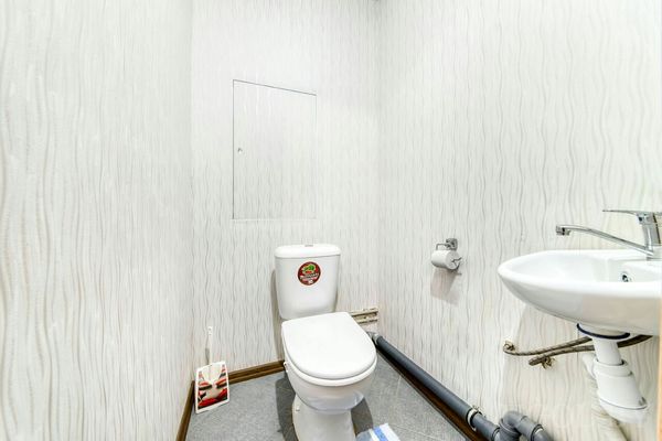 Узкое помещение с унитазом, умывальником, трубами, ковриком и декоративной плиткой.