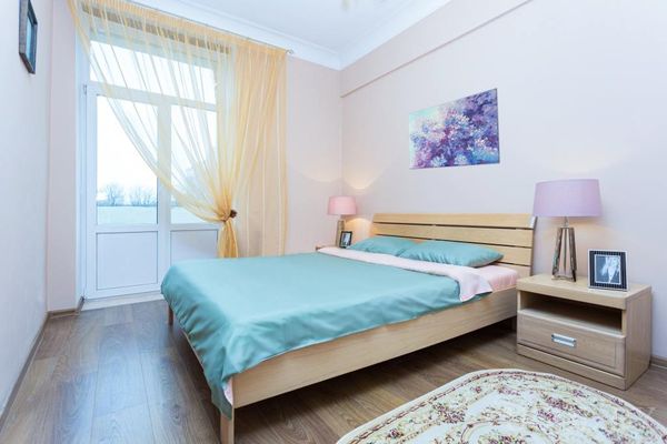 Светлая спальня с кроватью в бирюзовом покрывале, розовыми лампами на тумбочках и большим окном с жёлтыми шторами.