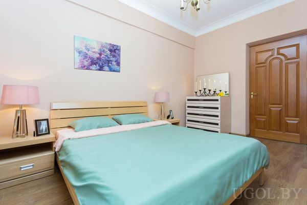 Уютная спальня с кроватью в мятном покрывале, розовыми лампами, белым комодом с зеркалом и свечами.