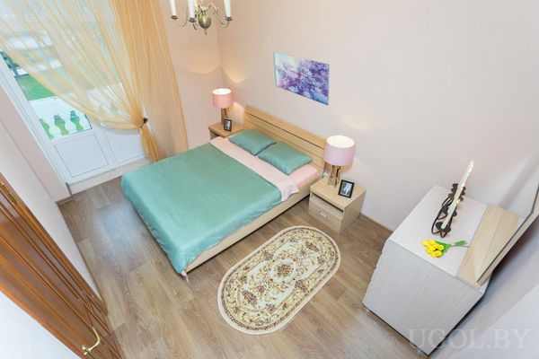 Комната с двуспальной кроватью, зелёным покрывалом, овальным ковром с узором и белым комодом с цветами.