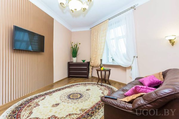 Комната с коричневым диваном с подушками, ковром с узором, столиком с фруктами и телевизором на стене.