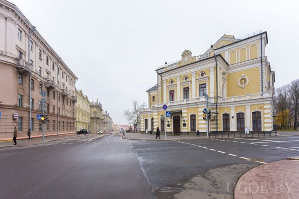 Городская улица с жёлтым историческим зданием с арочными окнами и балконом, рядом пешеходы.